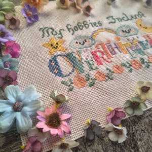 May include: A cross-stitch embroidery piece with the text "My Hobbies Include" and the word "CRAFTING" in colorful letters. The embroidery is surrounded by various artificial flowers in shades of pink, blue, and yellow.