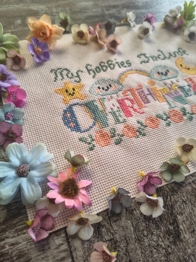 May include: A cross-stitch embroidery piece with the text "My Hobbies Include" and the word "CRAFTING" in colorful letters. The embroidery is surrounded by various artificial flowers in shades of pink, blue, and yellow.