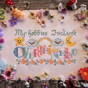 May include: Cross-stitch embroidery with the text "My hobbies include everything" in various colors. The design features a star, clouds, a rainbow, and a moon. The embroidery is surrounded by colorful artificial flowers and mushrooms.