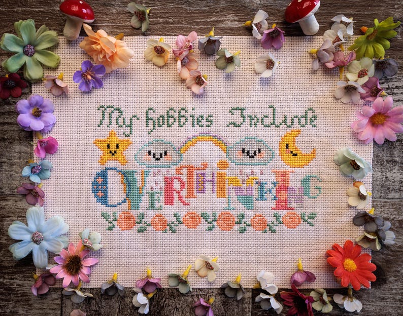 May include: Cross-stitch embroidery with the text "My hobbies include everything" in various colors. The design features a star, clouds, a rainbow, and a moon. The embroidery is surrounded by colorful artificial flowers and mushrooms.