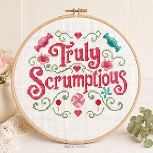 May include: A cross-stitch embroidery hoop with the words "Truly Scrumptious" in pink. The design includes pink candies, flowers, hearts, and green vines on a white background. The wooden hoop is round.