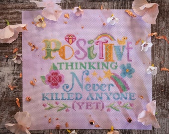 May include: A cross-stitch design with the text "POSITIVE THINKING NEVER KILLED ANYONE (YET)". The design features a rainbow, flowers, and stars, stitched on a pale purple fabric. The piece is surrounded by decorative elements.