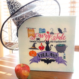 May include: A cross-stitch design with the text "Every Fairy Tale needs a good old-fashioned Villain" and Halloween-themed illustrations. A black witch hat with a spiderweb design and a red apple are also in the image.