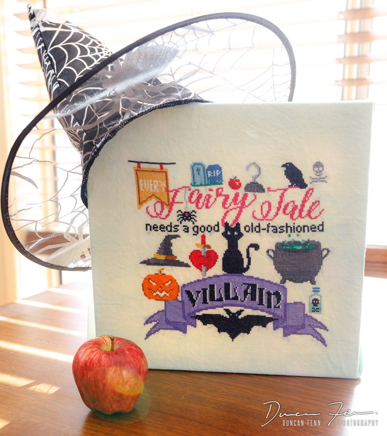 May include: A cross-stitch design with the text "Every Fairy Tale needs a good old-fashioned Villain" and Halloween-themed illustrations. A black witch hat with a spiderweb design and a red apple are also in the image.