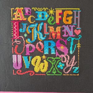 May include: A cross-stitch embroidery piece on black fabric featuring the alphabet in a variety of vibrant, multi-colored thread patterns. Each letter is uniquely styled with different colors, including pink, blue, yellow, orange, and green.