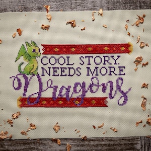 May include: Cross-stitch art on a light green fabric. The design features a green dragon, red and gold accents, and the text "COOL STORY. NEEDS MORE Dragons" in purple. Decorative gold flakes surround the artwork.