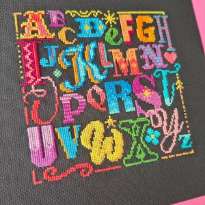 May include: A cross-stitch sampler on black fabric featuring the alphabet in a variety of vibrant, multi-colored thread styles. Each letter is uniquely designed with different patterns, including stars, hearts, and decorative flourishes in shades of pink, blue, yellow, green, and orange.