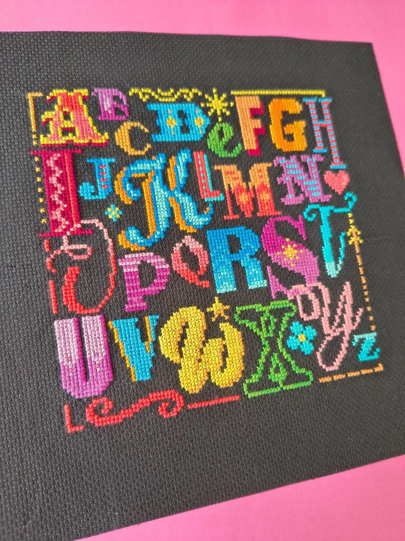 May include: A cross-stitch sampler on black fabric featuring the alphabet in a variety of vibrant, multi-colored thread styles. Each letter is uniquely designed with different patterns, including stars, hearts, and decorative flourishes in shades of pink, blue, yellow, green, and orange.