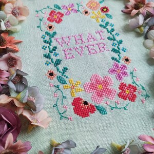 May include: Cross-stitch embroidery on light green fabric with the word "WHATEVER" in pink, surrounded by a floral wreath. The wreath includes pink, red, yellow, and purple flowers with green leaves. Decorative flowers frame the piece.