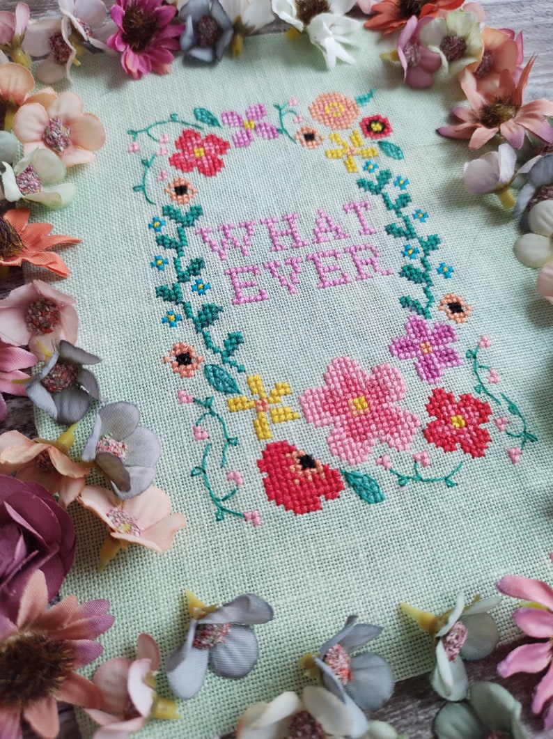 May include: Cross-stitch embroidery on light green fabric with the word "WHATEVER" in pink, surrounded by a floral wreath. The wreath includes pink, red, yellow, and purple flowers with green leaves. Decorative flowers frame the piece.
