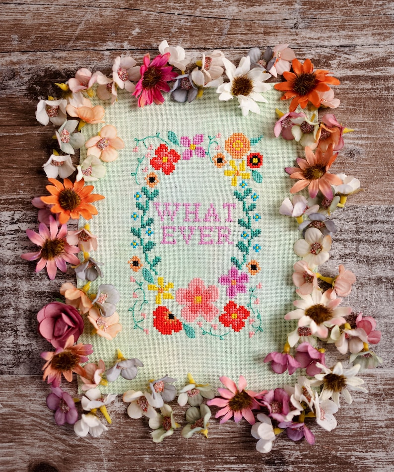 May include: A cross-stitch piece with the words "WHAT EVER" inside a floral wreath. The piece is surrounded by a border of colorful artificial flowers in shades of pink, orange, and white, set against a light green background.