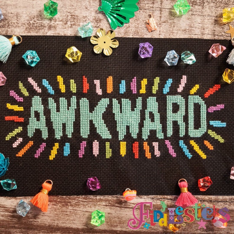 May include: A cross-stitch pattern with the word "AWKWARD" in teal surrounded by a colorful border of rainbow-colored stitches.