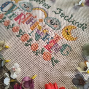 May include: A cross-stitch design on a cream-colored fabric, featuring the text "My Hobbies Include" and "Overthinking" in various colors. The design includes a star, clouds, a rainbow, and a crescent moon. Floral accents and decorative flowers surround the design.