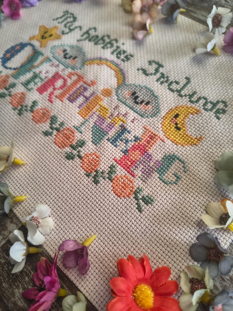 May include: A cross-stitch design on a cream-colored fabric, featuring the text "My Hobbies Include" and "Overthinking" in various colors. The design includes a star, clouds, a rainbow, and a crescent moon. Floral accents and decorative flowers surround the design.