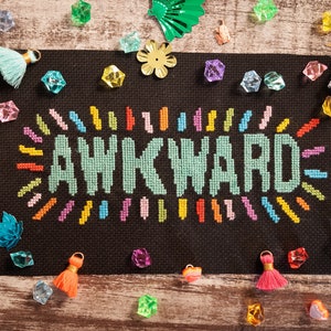 May include: A black canvas with the word "AWKWARD" stitched in colorful thread. The canvas is surrounded by colorful beads and tassels.
