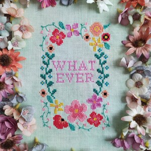 May include: Cross-stitch art piece with the word "WHATEVER" surrounded by a floral wreath in pink, red, yellow, and blue. The piece is on a light green background and framed by colorful artificial flowers.