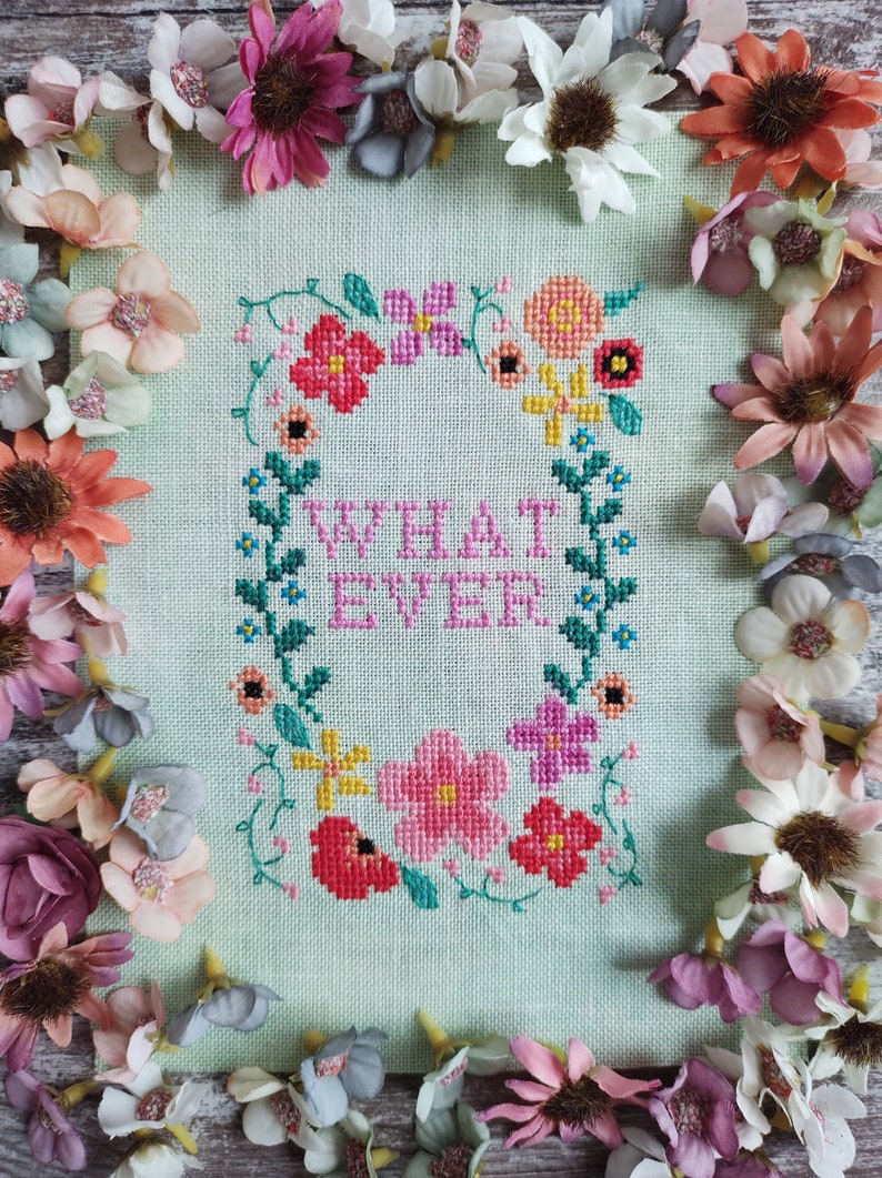 May include: Cross-stitch art piece with the word "WHATEVER" surrounded by a floral wreath in pink, red, yellow, and blue. The piece is on a light green background and framed by colorful artificial flowers.