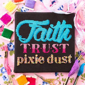 May include: Cross stitch embroidery on black fabric with the words "Faith Trust Pixie Dust" in colorful thread. The embroidery is surrounded by colorful embroidery floss spools and pink floral fabric.