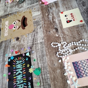 May include: A collection of colorful cross-stitch embroidery projects, including a portrait of a cartoon character, a mermaid tail, and the words "Awkward" and "I love you so much, right?"  The projects are displayed on a wooden surface with various decorative items, including beads, shells, and a string of pearls.