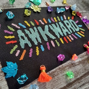 May include: A black canvas with the word "AWKWARD" stitched in colorful thread. The word is surrounded by a colorful, radiating pattern. The canvas is surrounded by colorful plastic gems.