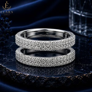 May include: Two silver-toned rings stacked vertically, each featuring a pavé setting with numerous small, clear, round-cut stones covering the entire outer surface of the bands. The rings have a polished metallic finish and a uniform, circular shape.