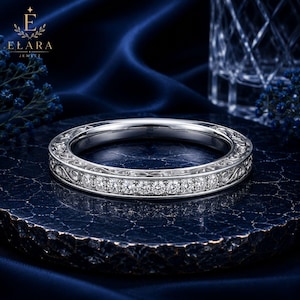 Könnte beinhalten: Ein silberfarbener Bandring mit einer zentralen Reihe kleiner, runder, klarer Steine. Die Seiten des Rings sind mit komplizierten, geschwungenen Metall-Filigranmustern verziert, die sich über den gesamten Umfang des Rings erstrecken.