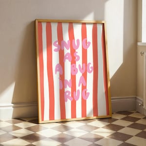 Può includere: Una stampa artistica da parete rettangolare in una cornice di legno chiaro con strisce verticali rosse e bianche. Il testo SNUG AS A BUG IN A RUG è stampato in uno stile disegnato a mano in rosa chiaro, centrato sul motivo a strisce.