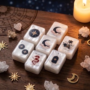 Può includere: Un set di sette tessere rettangolari bianche con finitura perlata, caratterizzate da vari disegni stampati tra cui stelle, una luna crescente, un drago rosso, una clessidra e il testo Flower e Dragon, disposte su una superficie in legno scuro.