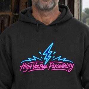 May include: A black hooded sweatshirt featuring a graphic print of a bright blue lightning bolt above the text High Voltage Personality written in a neon pink script font. The hoodie includes a front kangaroo pocket and matching drawstrings.