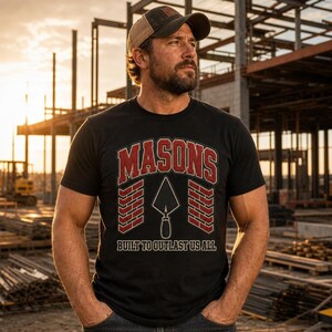 May include: A black t-shirt featuring a graphic with the word MASONS in red arched text above a white outline of a masonry trowel. Two red brick patterns flank the trowel, and the text BUILT TO OUTLAST US ALL is printed in white below.