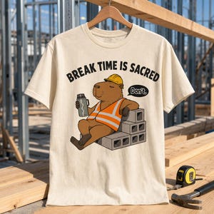 May include: A cream-colored t-shirt featuring a graphic of a brown capybara wearing a yellow hard hat and an orange safety vest. The capybara sits on gray concrete blocks holding a thermos. Text reads: BREAK TIME IS SACRED and Don't.