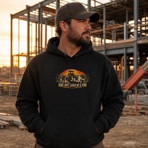 May include: A black hooded sweatshirt featuring a graphic print of a construction worker and a paving machine with the text "ONE HOT LOAD AT A TIME" in yellow and orange tones. The hoodie includes a front kangaroo pocket and long sleeves.