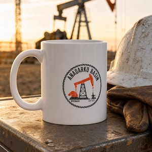 May include: A white ceramic mug featuring a circular graphic with a rope border. The design includes an orange oil pump jack, the text ANADARKO BASIN in black, and the words WOODWARD and ELK CITY printed below the pump jack illustration.