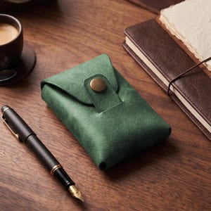 Könnte beinhalten: Ein rechteckiges, waldgrünes Etui mit strukturierter Oberfläche, das mit einer Umschlagklappe und einem einzelnen runden, messingfarbenen Druckknopf verschlossen wird. Das Etui hat ein strukturiertes Design mit sichtbaren Nähten an den Kanten.
