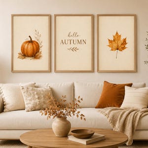 May include: Three framed wall art prints featuring a pumpkin on the left, the text hello AUTUMN in the center, and a maple leaf on the right. Each print has a light beige background and is enclosed in a simple light wood frame.