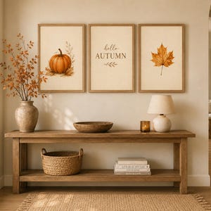 May include: Three framed wall art prints featuring a pumpkin, the text hello AUTUMN, and a maple leaf. The prints are displayed above a wooden console table with a vase of branches, a wooden bowl, a lamp, a candle, a basket, and stacked books.