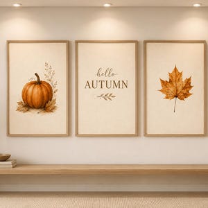 May include: A set of three framed wall art prints with light-colored backgrounds. The left print features an orange pumpkin, the center print displays the text "hello AUTUMN" with leaf accents, and the right print shows a single brown maple leaf.