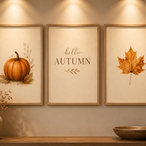 May include: A set of three framed wall art prints with light wood frames. The left print features an orange pumpkin with dried foliage. The centre print displays the text hello AUTUMN in brown. The right print shows a single brown maple leaf.