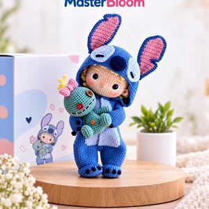 Peut inclure: Une poupée en crochet portant une combinaison à capuche bleue et bleu clair avec de grandes oreilles, tenant un petit jouet en peluche sarcelle avec un nœud rose et des yeux en bouton. Le texte MasterBloom est visible en haut.