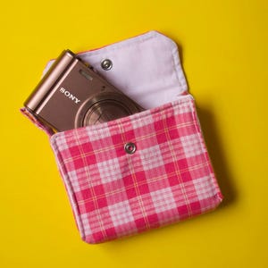 May include: A metallic bronze Sony digital camera partially tucked inside a small, rectangular fabric pouch featuring a pink and white plaid pattern with a silver metal snap button closure on the front flap and a white interior lining.
