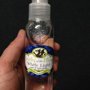 Pode incluir: Uma garrafa de vidro transparente com um bico de pulveriza&ccedil;&atilde;o branco e um r&oacute;tulo que diz "Lady of the Moss White Light aura & room cleanse". O r&oacute;tulo apresenta um design de drag&atilde;o verde e azul.