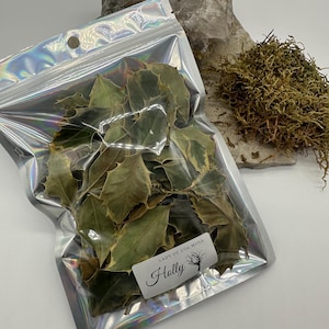Puede incluir: Una bolsa holográfica transparente llena de hojas de acebo secas. La bolsa tiene una pequeña etiqueta que dice "Lady of the Moss" y "Holly". Una gran roca de color claro y un montón de musgo seco están en el fondo.
