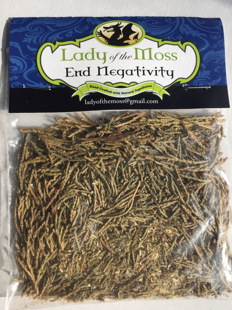 Puede incluir: Una bolsa de pl&aacute;stico transparente llena de agujas de cedro secas de color verde y marr&oacute;n. La bolsa tiene una etiqueta que dice "Lady of the Moss End Negativity" con un logotipo de drag&oacute;n y el texto "Hand Crafted with Natural Ingredients" y "ladyofthemoss@gmail.com".