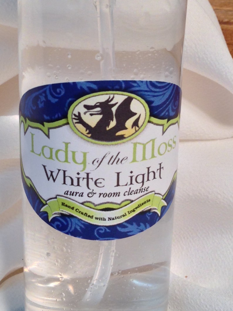 Pode incluir: Uma garrafa de vidro transparente com um r&oacute;tulo azul e verde que diz "Lady of the Moss White Light aura & room cleanse Hand Crafted with Natural Ingredients". O r&oacute;tulo apresenta um drag&atilde;o verde sobre um fundo amarelo.