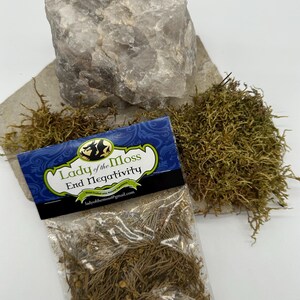Puede incluir: Una peque&ntilde;a bolsa de pl&aacute;stico transparente con una etiqueta que dice "Lady of the Moss End Negativity" y contiene musgo seco. La bolsa est&aacute; sobre un trozo de papel marr&oacute;n con un gran cristal de cuarzo transparente y un mont&oacute;n de musgo verde.