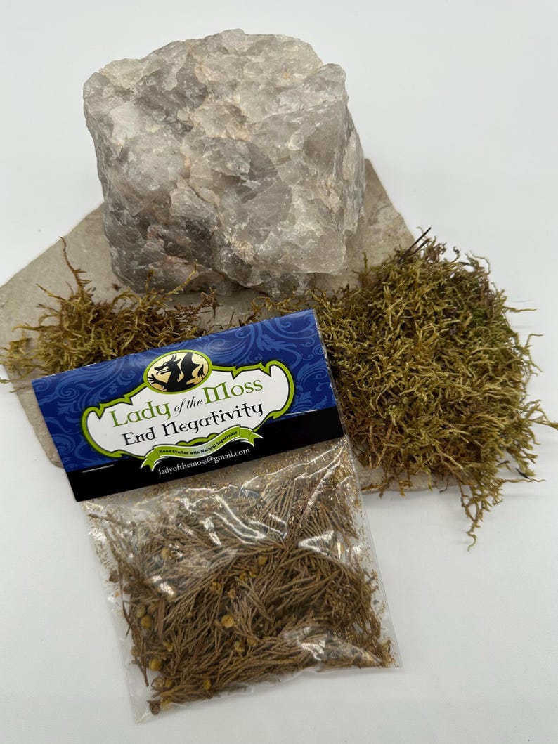 Puede incluir: Una peque&ntilde;a bolsa de pl&aacute;stico transparente con una etiqueta que dice "Lady of the Moss End Negativity" y contiene musgo seco. La bolsa est&aacute; sobre un trozo de papel marr&oacute;n con un gran cristal de cuarzo transparente y un mont&oacute;n de musgo verde.