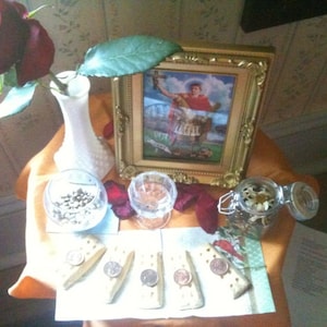 May include: A still life arrangement featuring a deep red rose in a white vase, a framed religious image, small glasses with contents, crackers with coins, and a jar with dried flowers. The items are arranged on a table with an orange cloth.