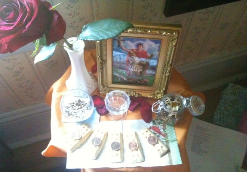 Puede incluir: Un bodeg&oacute;n con una rosa roja oscura en un jarr&oacute;n blanco, una imagen religiosa enmarcada, vasos peque&ntilde;os con contenido, galletas saladas con monedas y un tarro con flores secas. Los elementos est&aacute;n dispuestos sobre una mesa con un pa&ntilde;o naranja.