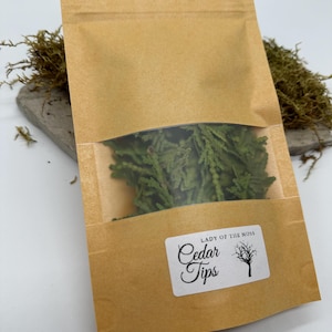 Może przedstawiać: Brązowa papierowa torba z przezroczystym okienkiem, wypełniona zielonymi końc&oacute;wkami cedru. Torba ma białą etykietę z napisem "Lady of the Moss" i "Cedar Tips" z ilustracją drzewa. Torba leży na szarym kamieniu.