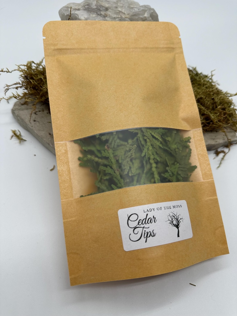 Może przedstawiać: Brązowa papierowa torba z przezroczystym okienkiem, wypełniona zielonymi końc&oacute;wkami cedru. Torba ma białą etykietę z napisem "Lady of the Moss" i "Cedar Tips" z ilustracją drzewa. Torba leży na szarym kamieniu.